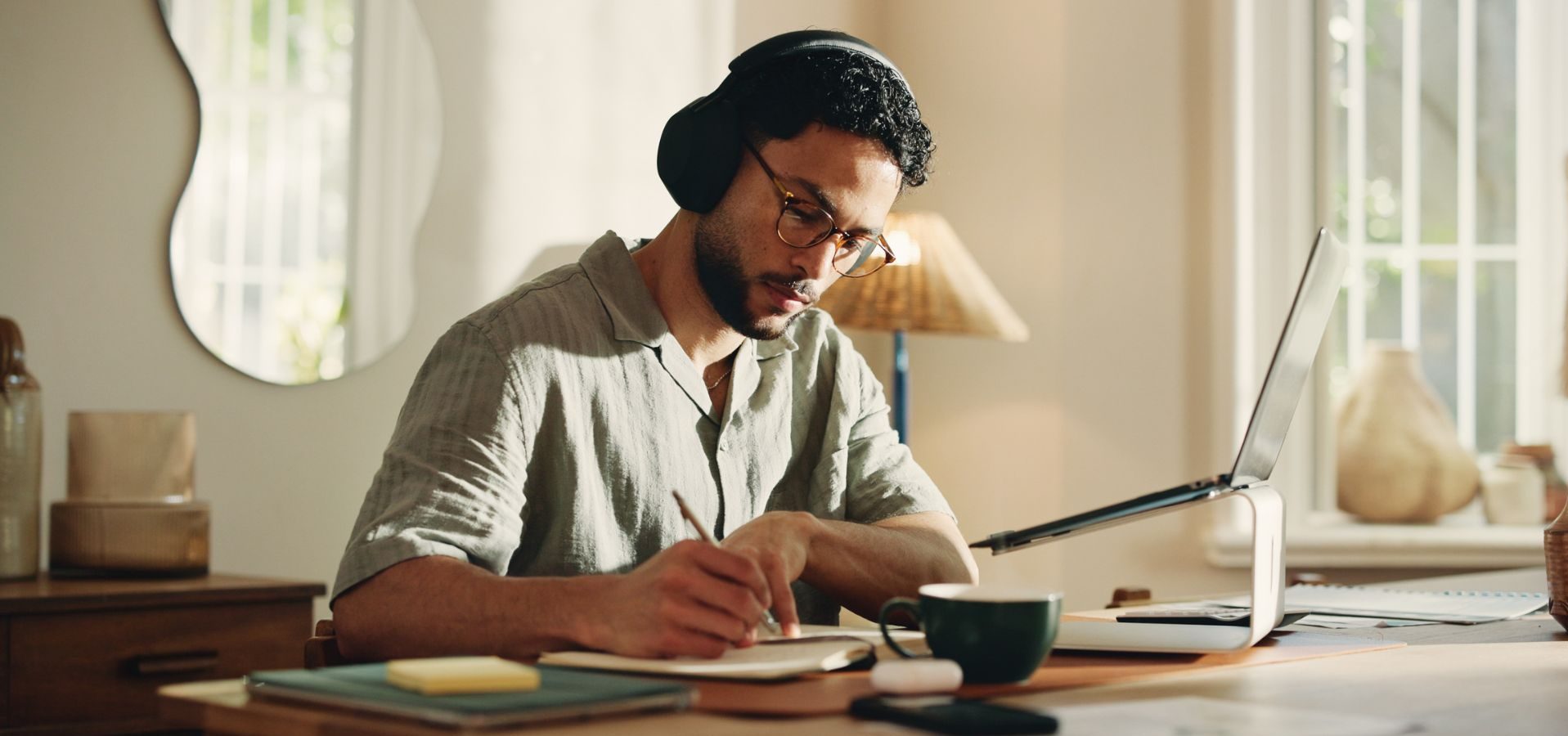 Man wearing headphones writes in a notebook beside his laptop
