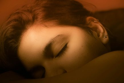 A close-up of a person with brown hair sleeping on their side, eyes closed, with warm, soft orange lighting highlighting their face and ear—perfect inspiration for quality sleep tips.