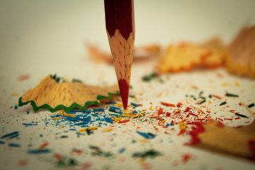 A sharpened red colored pencil points downward amid colorful pencil shavings and scattered fragments of blue, red, and yellow pencil on a white surface.