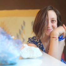 A young woman with short brown hair smiles while resting her head on her hand, wearing a blue patterned shirt and a red string bracelet. Crystals and stones are on the table, suggesting she’s ready for a Tarot Reading.