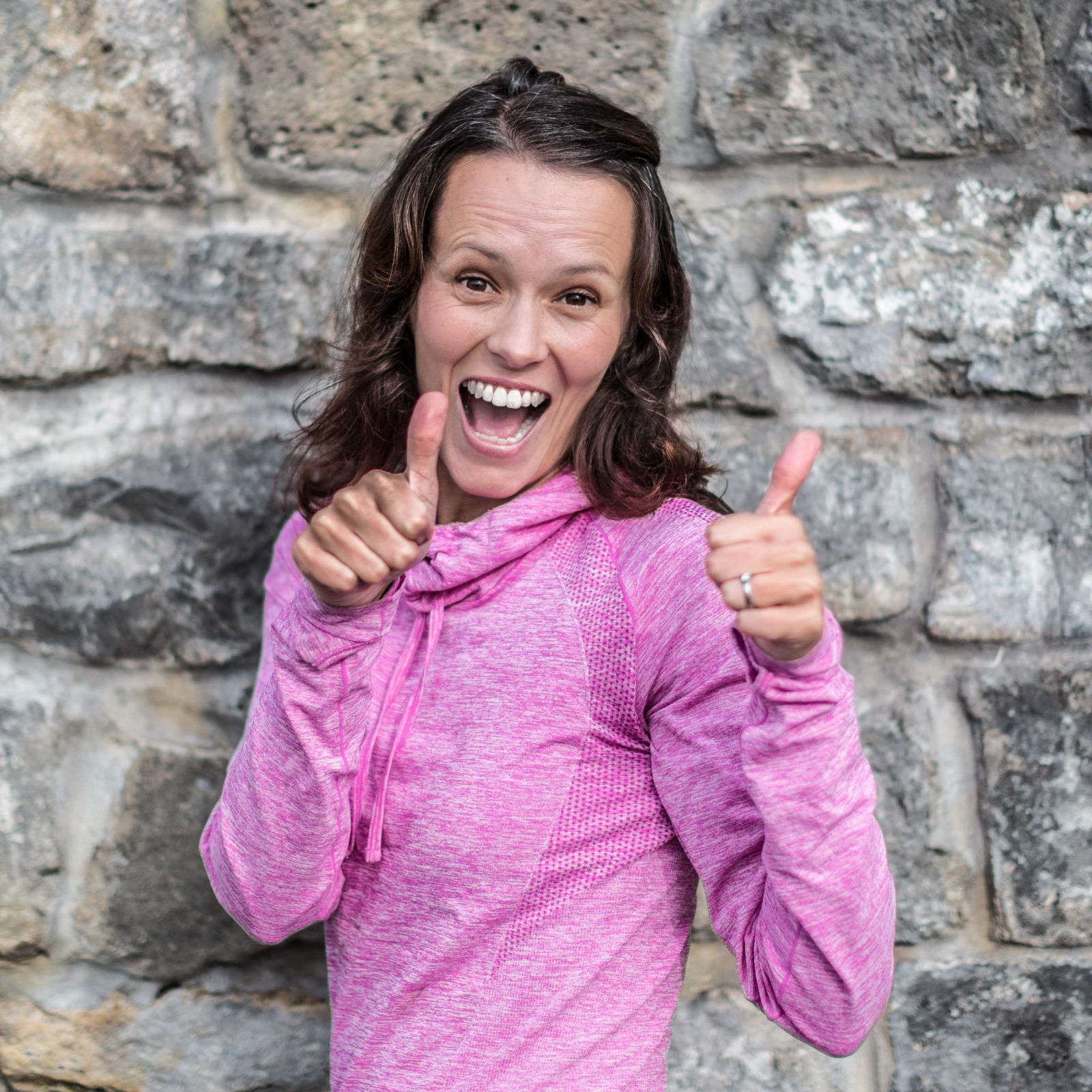 A woman in a pink athletic hoodie smiles and gives two thumbs up in front of a stone wall, celebrating progress with the ADHD Coach for Overwhelm, Focus & Calm.