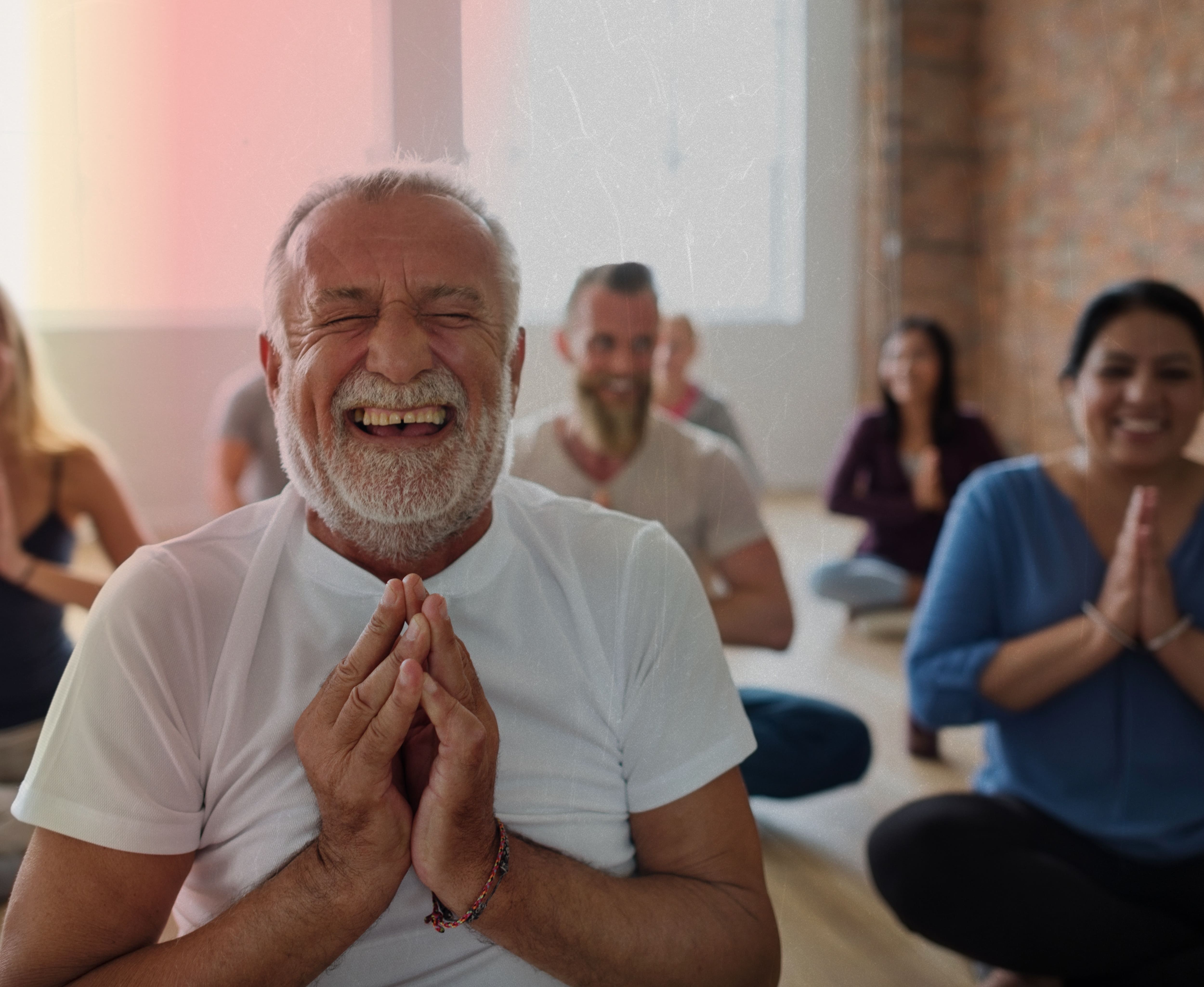 Participants in the Virtual Laughter Yoga Workshop for Team Building sit cross-legged, smiling and holding a prayer pose. An older man leads with joyful laughter, creating an uplifting online experience for everyone.
