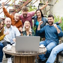 A group of excited people gather outdoors, cheering and raising their fists as they participate in the Virtual Happiness Workshop for Team Building, watching a laptop screen together during a fun online team event.