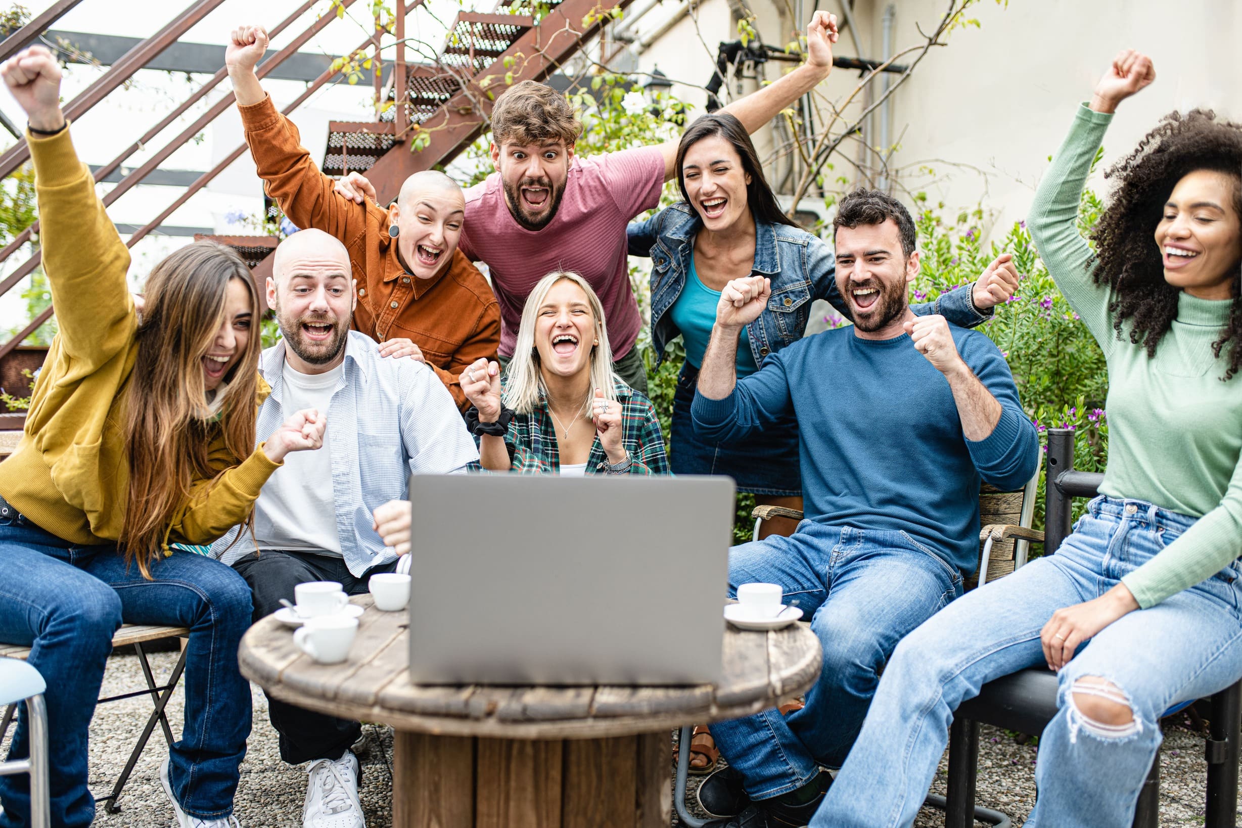 A group of excited people gather outdoors, cheering and raising their fists as they participate in the Virtual Happiness Workshop for Team Building, watching a laptop screen together during a fun online team event.