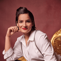 A person with short dark hair and red lipstick, dressed in a white shirt and suspenders, sits smiling on a gold chair against a reddish-brown background, representing the Virtual Happiness Workshop for Team Building.