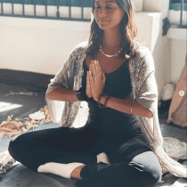 A woman sits cross-legged on a mat indoors, eyes closed, with hands in a prayer position at her chest. She wears a light shawl, dark clothes, and white socks, appearing calm and meditative in natural sunlight.