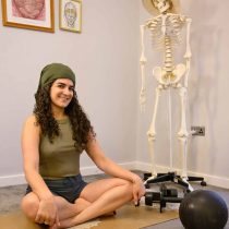 A smiling woman with curly hair and a green headscarf sits cross-legged on a yoga mat indoors. Next to her is a human skeleton model wearing a hat. Exercise equipment and framed art are visible in the background.