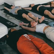 Four women lie on yoga mats indoors with their arms extended above their heads, relaxing in a restful pose. They appear calm and peaceful, wearing comfortable workout clothes.