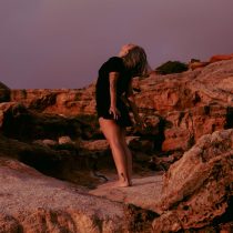 A person in a black dress stands barefoot on rocky terrain, leaning back with their arms extended and head tilted upward, surrounded by reddish-brown rocks under a purple-tinged sky.