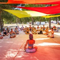 A group of people sit cross-legged in a circle on a mat outdoors, participating in a guided meditation or yoga session under colorful fabric canopies, surrounded by trees.