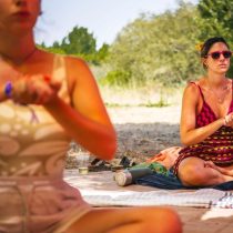 Two women sit cross-legged outdoors on blankets, one in focus wearing sunglasses and a patterned dress, the other blurred in the foreground, both appearing to participate in a meditative or relaxing activity.