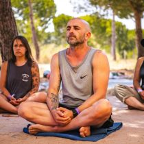 A man with a shaved head and tattoos sits cross-legged on a mat outdoors, eyes closed in meditation. Two women sit nearby, also meditating. Trees and sunlight are visible in the background.