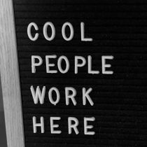 A black felt letter board with white plastic letters spelling “COOL PEOPLE WORK HERE,” positioned vertically. A wooden frame is visible on the left side of the board.