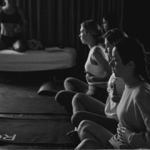 Black and white photo of four women sitting on yoga mats in a row, eyes closed and hands on their stomachs, practicing deep breathing in a calm indoor space with curtains in the background.