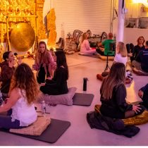 A group of people sit in pairs on cushions and mats in a spacious room with golden statues and a gong, engaging in face-to-face conversation or meditation exercises. The atmosphere appears calm and focused.