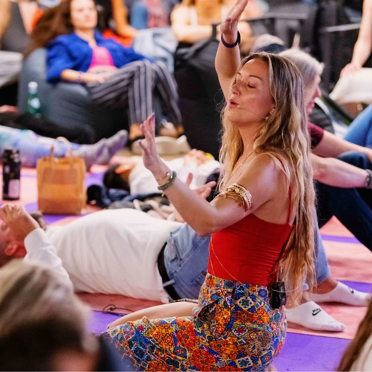 A woman with long blonde hair, wearing a red top and patterned skirt, sits on the floor with eyes closed and hands raised, appearing to meditate or take part in a group activity with others sitting and lying nearby.