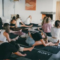 A group of women sit on yoga mats in a bright room, stretching and doing yoga exercises together. Some are leaning forward with legs extended, and the mats are labeled ROXY. A fireplace and artwork are visible in the background.