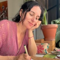 A woman with dark curly hair, wearing a pink knitted top, sits at a table writing with a pen. She rests her chin on her hand and smiles softly. A potted plant and embroidered bag are visible in the background.