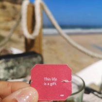 A hand holds a pink tea tag that says, This life is a gift, with a blurry beach scene, rope barrier, and blue sky in the background.