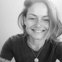 A person with shoulder-length hair smiles at the camera, wearing a dark t-shirt and a necklace with a pendant. The photo is black and white, and their arm shows a tattoo. The background is plain.