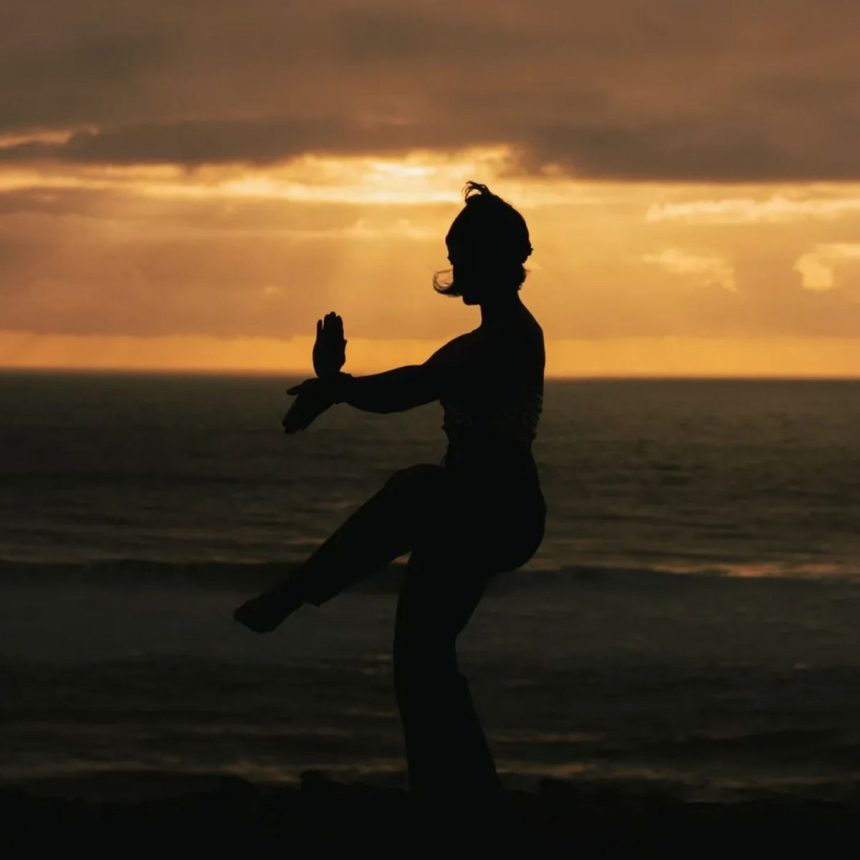 Silhouette of a person practicing martial arts or tai chi on a beach at sunset, with the ocean and dramatic orange sky in the background.
