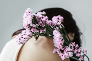A person in a sleeveless white top sits with their head bowed, holding a bouquet of small pink flowers over their shoulder against a soft gray background.
