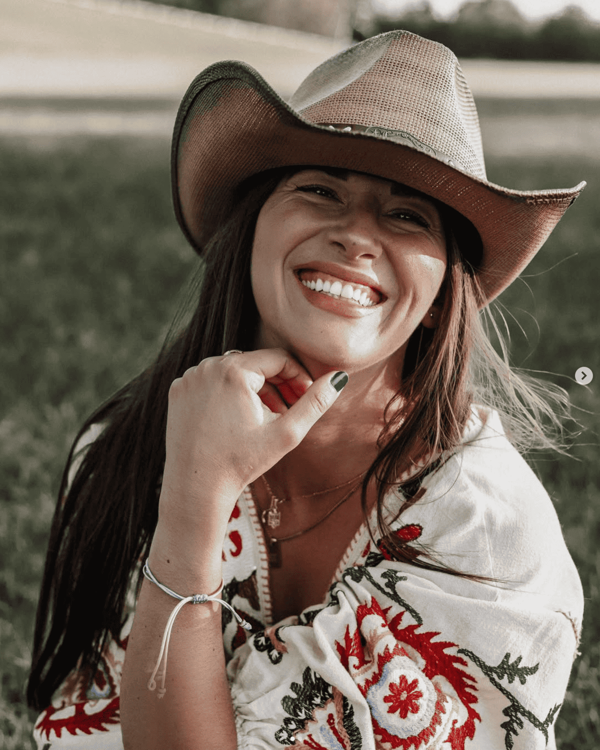 A woman wearing a cowboy hat and a patterned shawl smiles brightly while sitting on grass, with her hand resting near her chin. The background is blurred, highlighting her joyful expression.