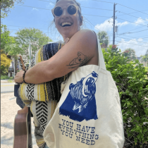 A smiling woman wearing sunglasses holds a ukulele and a striped blanket. She carries a tote bag with a blue bear illustration and the words You have everything you need. Trees and power lines are visible in the background.