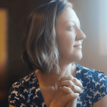A woman with short brown hair, wearing a blue and white dress, sits clasping her hands and gazes thoughtfully toward light—reflecting on insights from the "Messaging Magic: Your Soul-Words Tarot Reading.