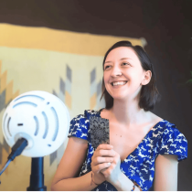 A smiling woman in a blue patterned dress holds tarot cards before a microphone, ready to deliver a "Messaging Magic: Your Soul-Words Tarot Reading" in her serene, tapestry-adorned space accented by a green plant.