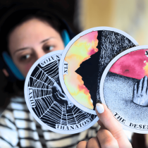 A person with light skin, dark hair, headphones, and a striped shirt holds up three illustrated tarot cards—highlighting "Messaging Magic: Your Soul-Words Tarot Reading," a session for intuitive business guidance. The focus is on the cards, not the face.