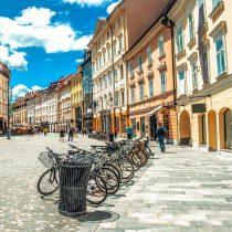 A row of bicycles is parked along a cobblestone street lined with colorful historic buildings under a bright blue sky with scattered clouds. Pedestrians walk nearby, enjoying the sunny day.
