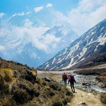 Two hikers with backpacks walk along a mountain trail beside a small stream, surrounded by rocky terrain and patches of snow, with tall, snowy peaks and clouds in the background.