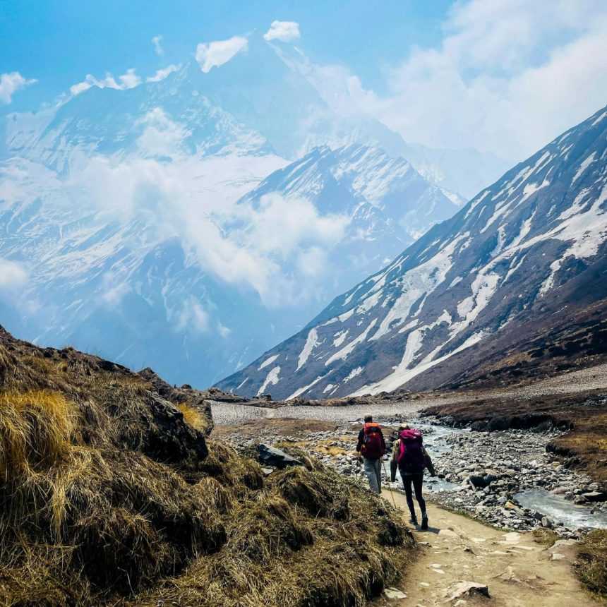Two hikers with backpacks walk along a mountain trail beside a small stream, surrounded by rocky terrain and patches of snow, with tall, snowy peaks and clouds in the background.