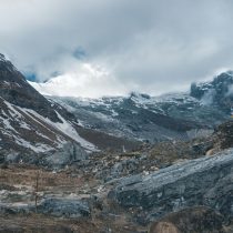 A rocky mountain landscape with patches of snow, cloudy skies, and rugged terrain. Snow-capped peaks are visible in the background, while the foreground features rocks and sparse vegetation.