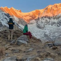 Two people in winter clothing sit and stand on rocky ground, looking at snow-capped mountains glowing orange in the sunrise or sunset. The landscape is rugged with dry grass and dramatic peaks.