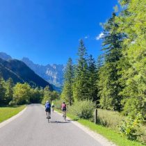 Two cyclists ride on a paved road amid tall green trees and mountains under a clear blue sky—an idyllic setting, much like the Adventure to Annapurna Base Camp Hiking Tour experience.