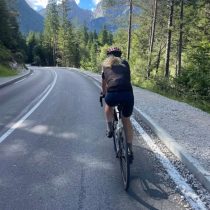 A cyclist in a helmet rides up a winding mountain road amid tall trees and distant peaks under a clear blue sky, evoking the spirit of the Adventure to Annapurna Base Camp Hiking Tour.