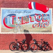 A vibrant mural on a red brick wall reads “Greetings from Clinton, MO” with local sights and “#VisitClintonMo.” Two bikes stand before it, their shadows hinting at journeys—like the Adventure to Annapurna Base Camp Hiking Tour.