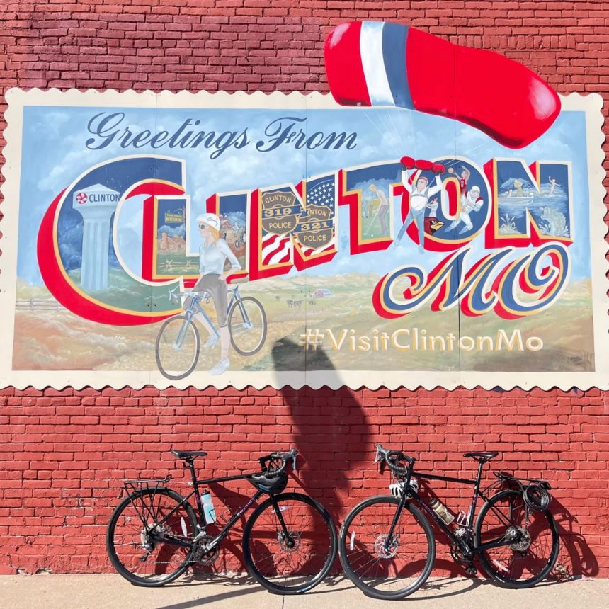 A vibrant mural on a red brick wall reads “Greetings from Clinton, MO” with local sights and “#VisitClintonMo.” Two bikes stand before it, their shadows hinting at journeys—like the Adventure to Annapurna Base Camp Hiking Tour.