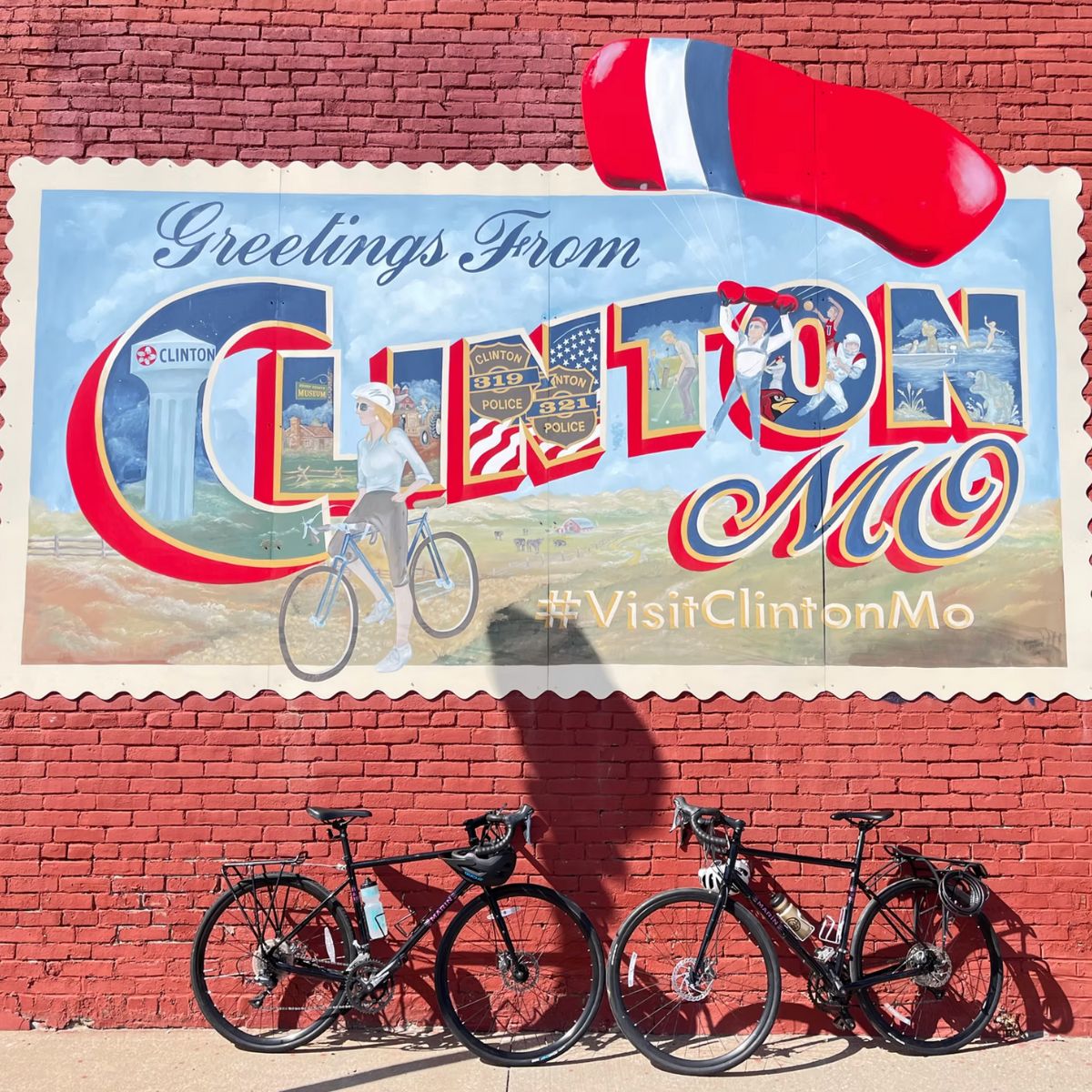 A vibrant mural on a red brick wall reads “Greetings from Clinton, MO” with local sights and “#VisitClintonMo.” Two bikes stand before it, their shadows hinting at journeys—like the Adventure to Annapurna Base Camp Hiking Tour.