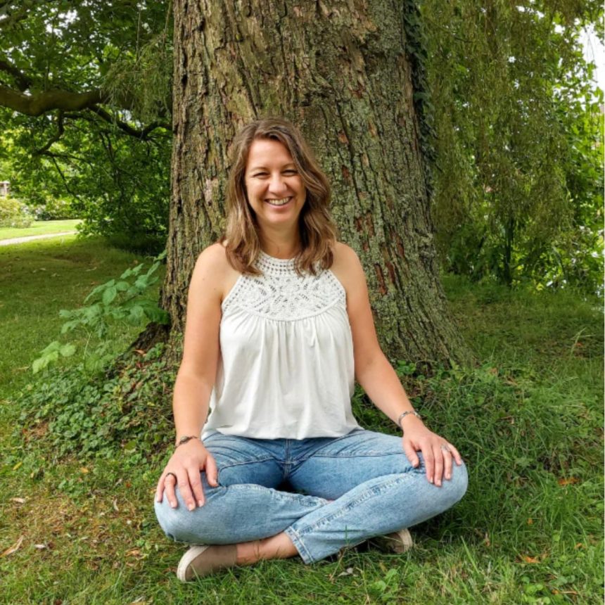 A woman with brown hair, wearing a white sleeveless top and blue jeans, sits cross-legged on grass in front of a large tree, smiling. Lush green foliage surrounds her in the background.