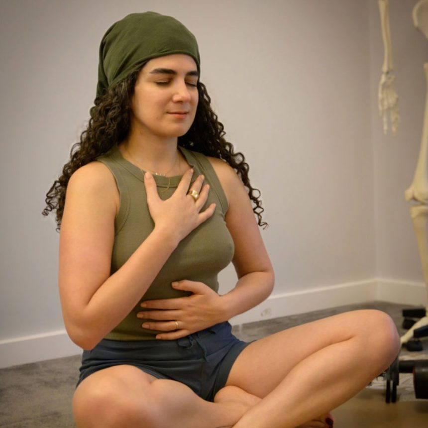 A woman with long curly hair, wearing a green headscarf and tank top, sits cross-legged on the floor with eyes closed, one hand on her chest and the other on her abdomen, practicing mindful breathing.