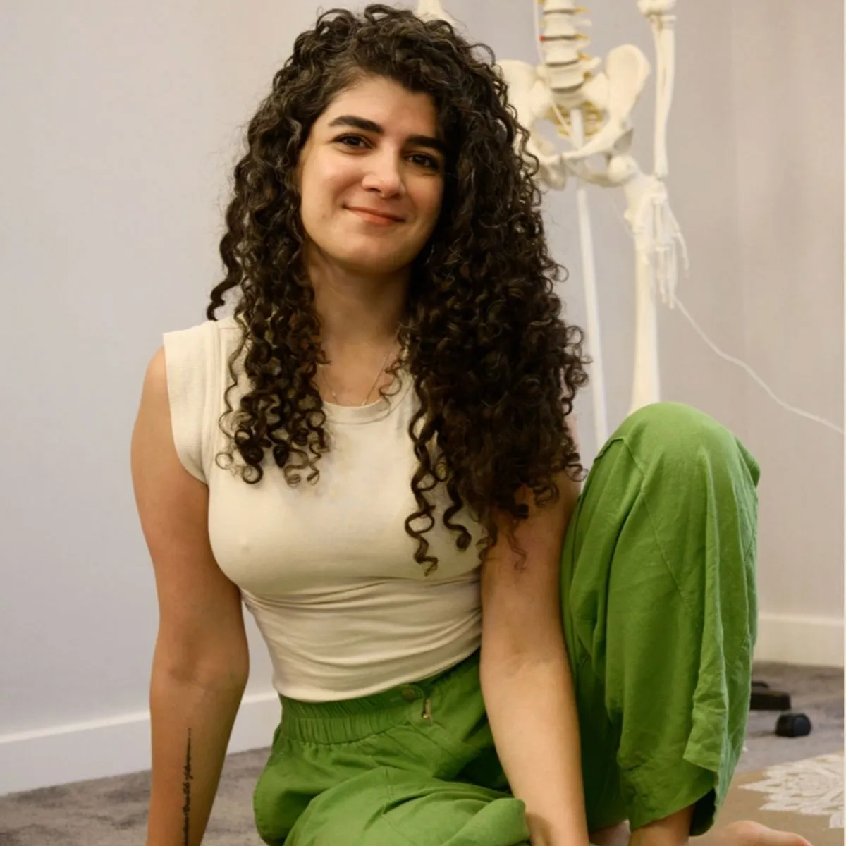 A woman in a sleeveless white top and green pants smiles at the camera while sitting on the floor. A skeleton model in the background highlights Body Check-In’s focus on postural imbalance and pain assessment themes.