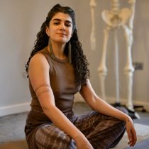 A person with long curly hair sits cross-legged indoors, wearing a sleeveless brown top and patterned brown pants. A skeleton model stands in the blurred background near a cream-colored wall.