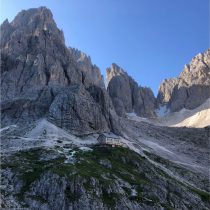 A mountain hut sits on a rocky, grassy slope at the base of steep, jagged peaks under a clear blue sky. Light and shadow highlight the rugged terrain and dramatic landscape.