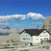 A mountain lodge with a dark roof and white walls sits on rocky terrain under a bright blue sky with scattered clouds. Distant mountains and cliffs are visible in the background.