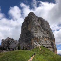 A group of people hike up a narrow dirt trail on a grassy hill toward a towering, rugged rock formation under a partly cloudy sky.