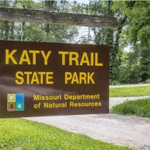 A brown sign reads Katy Trail State Park with Missouri Department of Natural Resources below, surrounded by green grass and trees.