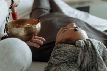 A person lying on a bed with eyes closed, a crystal on their forehead, and a practitioner holding a singing bowl near their head during a sound healing or meditation session.